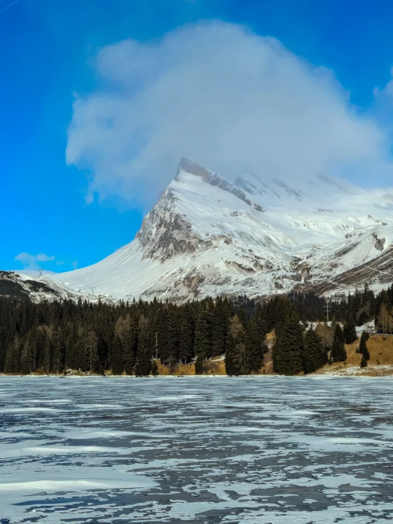 Winter landscape of frozen Lake Isola in San Bernardino, Swiss Alps, with a distinctive snow-covered mountain peak, a cloud resting on its summit, and a dense pine forest under a bright blue sky.